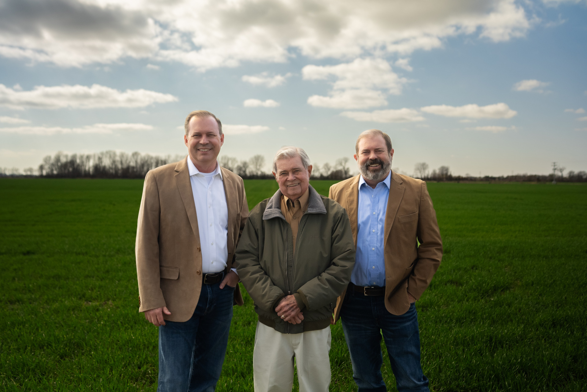 Three men stand in a green field under a cloudy sky. They are smiling, wearing casual jackets and jeans. Trees line the horizon.
